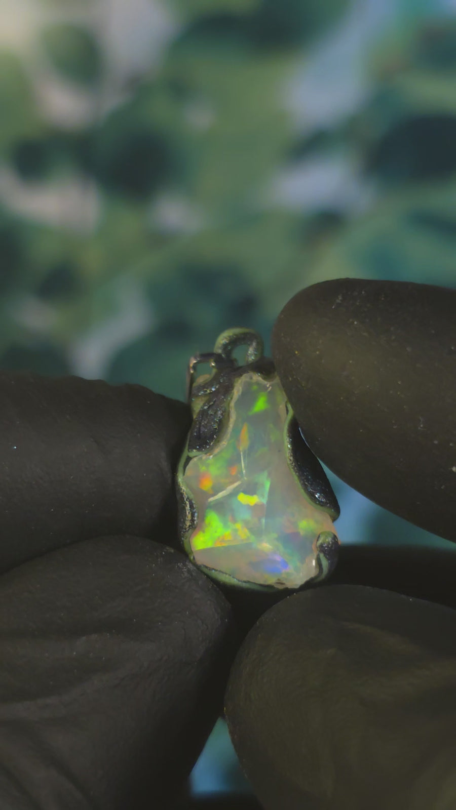 Video: raw uncut Ethiopian Welo opal pendant. Colour: green, blue-green, with red hints, vivid. Body: white. Held in a gloved hand against a green background.