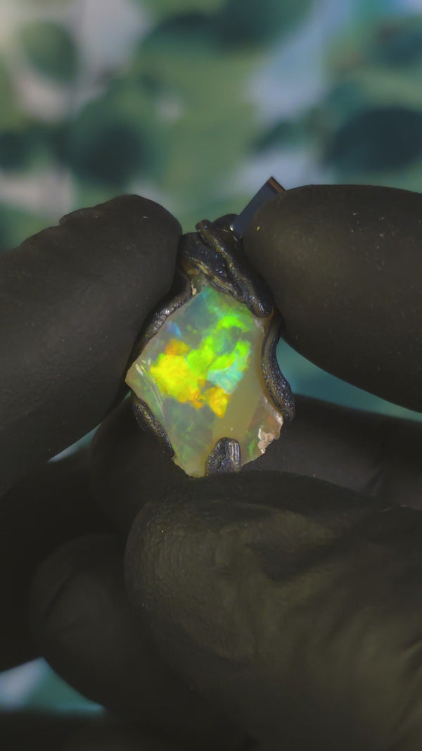 Video: raw uncut Ethiopian Welo opal pendant. Colour: blue, red, with green and yellow accents, vivid. Body: white. Held in a gloved hand against a green background.