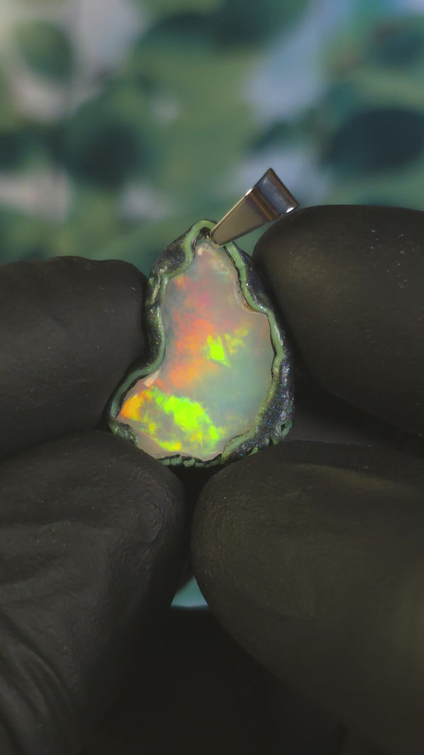 Video: raw uncut Ethiopian Welo opal pendant. Colour: full spectrum rainbow, moderate. Body: white. Held in a gloved hand against a green background.
