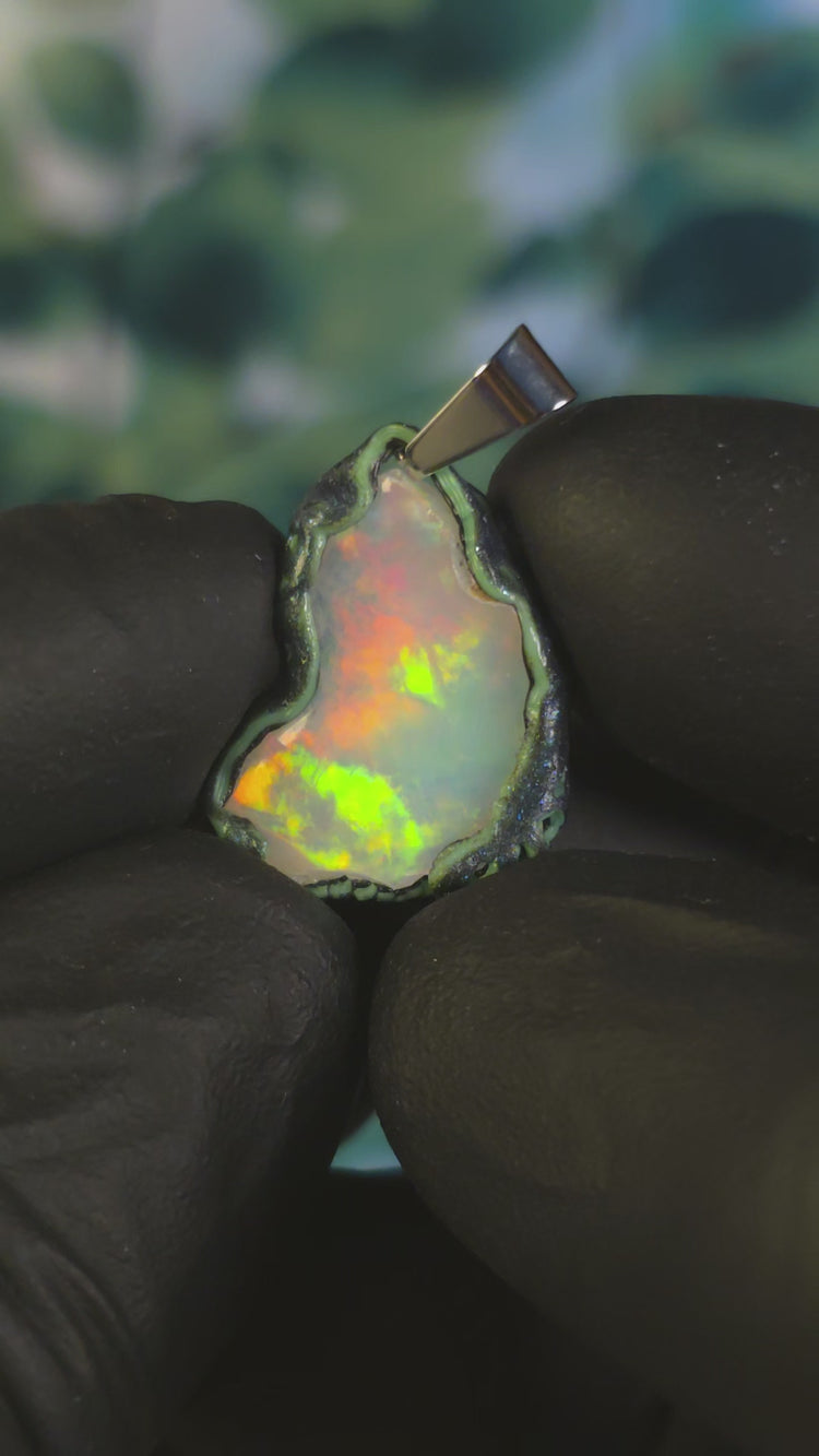 Video: raw uncut Ethiopian Welo opal pendant. Colour: full spectrum rainbow, moderate. Body: white. Held in a gloved hand against a green background.