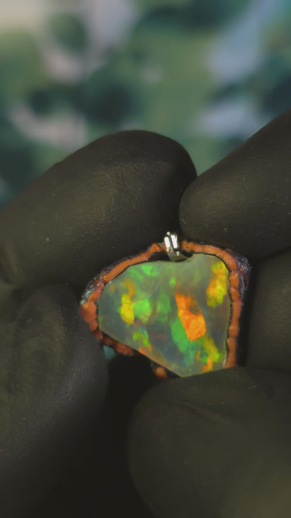 Video: raw uncut Ethiopian Welo opal pendant. Colour: full rainbow, vivid. Body: dark. Held in a gloved hand against a green background.