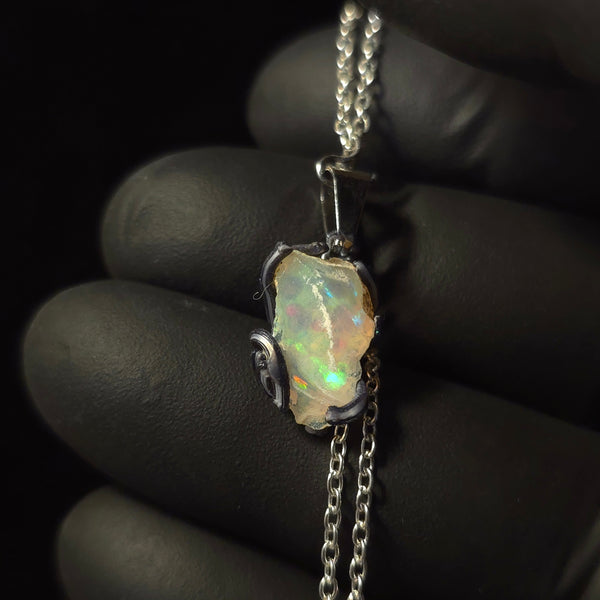 Raw uncut Ethiopian Welo opal pendant. Colour: green, moderate, comes and goes. Body: crystal clear. Held in a gloved hand against a dark background.