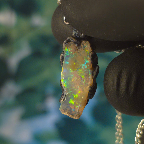 Raw uncut Ethiopian Welo opal pendant. Colour: blue and green, vivid. Body: transparent. Held in a gloved hand against a green background.