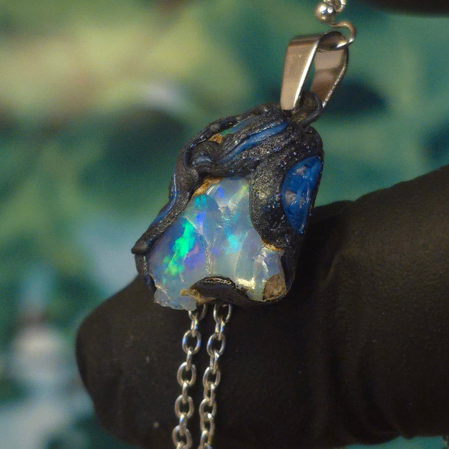 Raw uncut Ethiopian Welo opal pendant. Colour: green, blue, vivid. Body: blue. Held in a gloved hand against a green background.