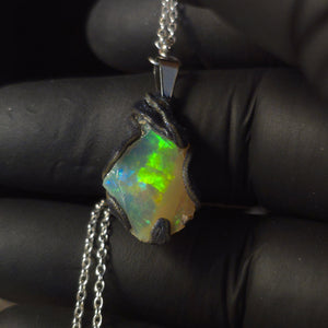 Raw uncut Ethiopian Welo opal pendant. Colour: blue, red, with green and yellow accents, vivid. Body: white. Held in a gloved hand against a dark background.