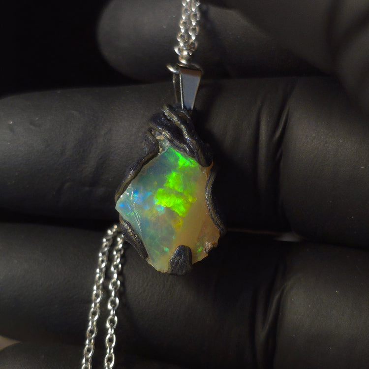 Raw uncut Ethiopian Welo opal pendant. Colour: blue, red, with green and yellow accents, vivid. Body: white. Held in a gloved hand against a dark background.