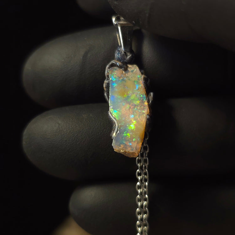 Raw uncut Ethiopian Welo opal pendant. Colour: blue and green, vivid. Body: transparent. Held in a gloved hand against a dark background.