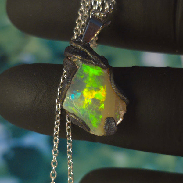 Raw uncut Ethiopian Welo opal pendant. Colour: blue, red, with green and yellow accents, vivid. Body: white. Held in a gloved hand against a green background.