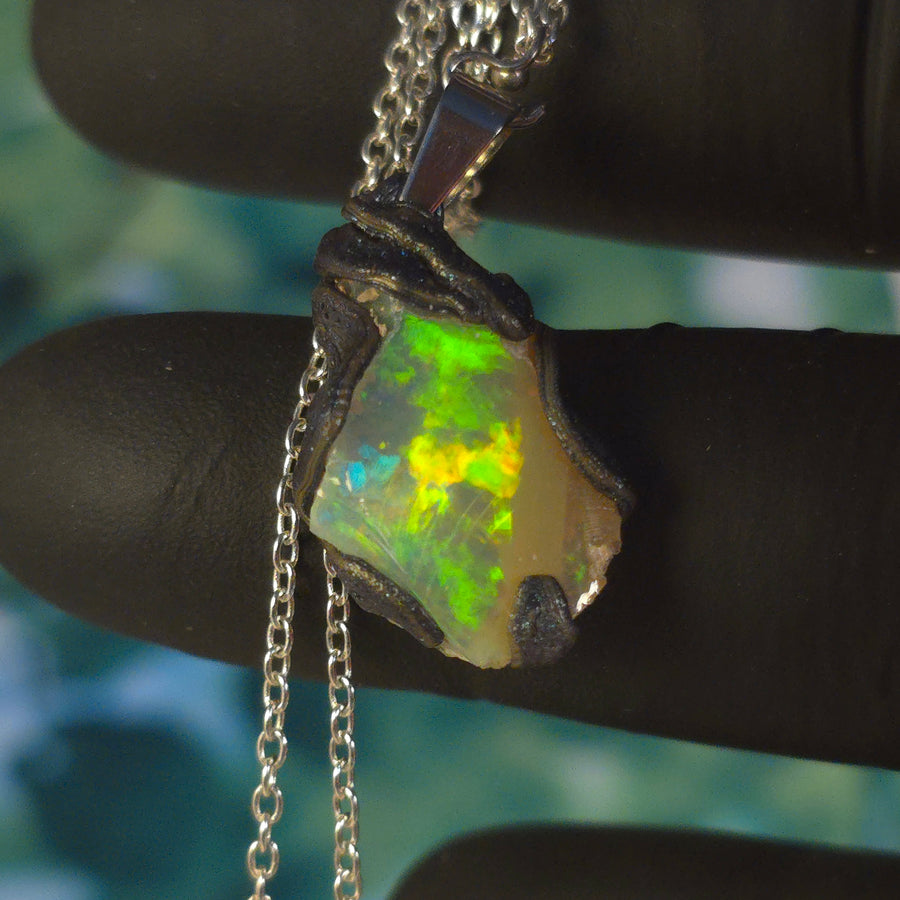 Raw uncut Ethiopian Welo opal pendant. Colour: blue, red, with green and yellow accents, vivid. Body: white. Held in a gloved hand against a green background.
