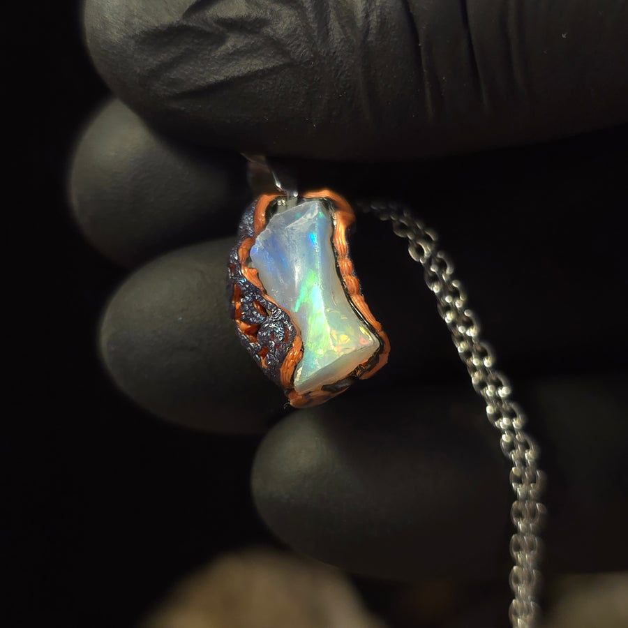 Raw uncut Ethiopian Welo opal pendant. Colour: red, gold with blue undertones, strong. Body: cloudy. Held in a gloved hand against a dark background.