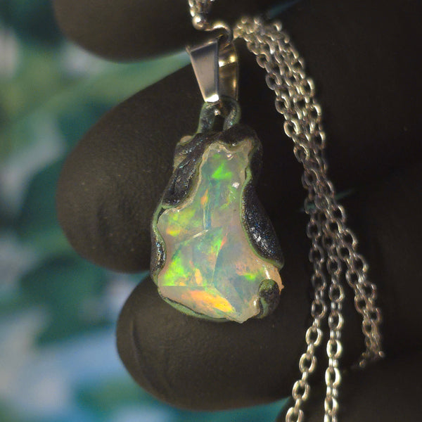 Raw uncut Ethiopian Welo opal pendant. Colour: green, blue-green, with red hints, vivid. Body: white. Held in a gloved hand against a green background.