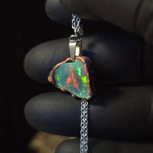 Raw uncut Ethiopian Welo opal pendant. Colour: full rainbow, vivid. Body: dark. Held in a gloved hand against a dark background.