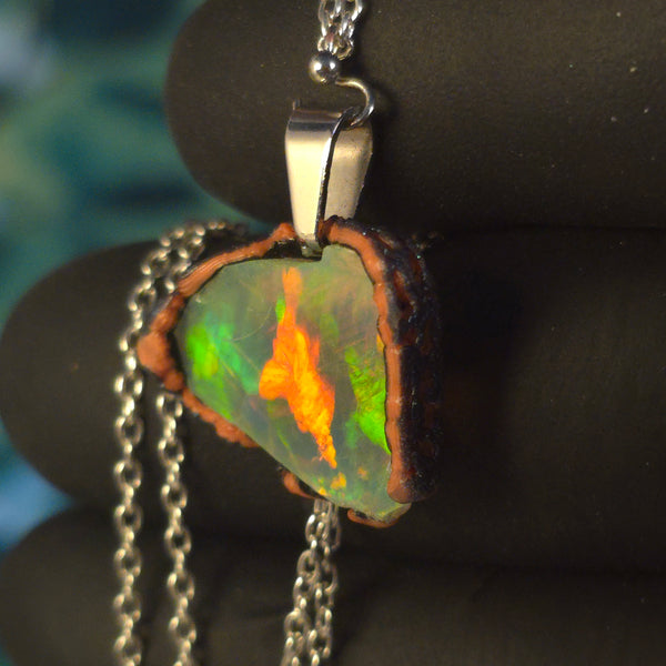 Raw uncut Ethiopian Welo opal pendant. Colour: full rainbow, vivid. Body: dark. Held in a gloved hand against a dark background.