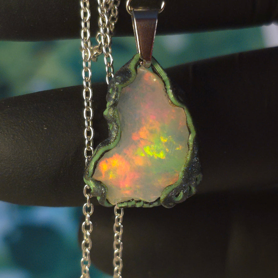 Raw uncut Ethiopian Welo opal pendant. Colour: full spectrum rainbow, moderate. Body: white. Held in a gloved hand against a green background.