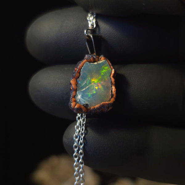 Raw uncut Ethiopian Welo opal pendant. Colour: red, green and gold, moderate. Body: clear with hazy surface. Held in a gloved hand against a dark background.