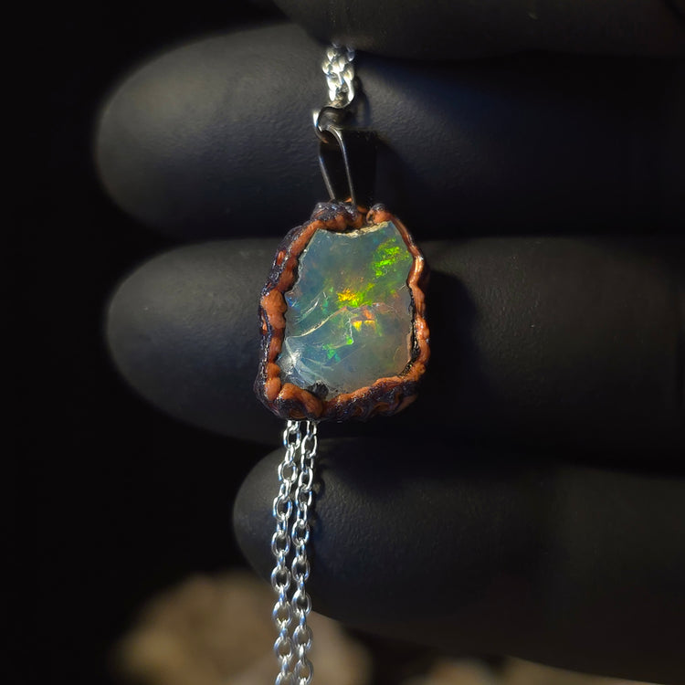 Raw uncut Ethiopian Welo opal pendant. Colour: red, green and gold, moderate. Body: clear with hazy surface. Held in a gloved hand against a dark background.